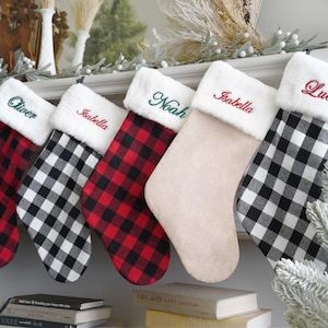 May include: Five Christmas stockings with white faux fur cuffs, each embroidered with a name: Oliver, Isabella, Noah, Isabella, and Lucas. The stockings feature red and black buffalo plaid, black and white buffalo plaid, and solid beige fabric.
