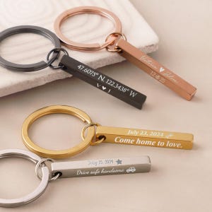 Peut inclure: Plusieurs porte-clés personnalisés de différentes couleurs métalliques. L'un est noir avec des coordonnées, un autre est en or rose avec des noms et une date, et d'autres ont des dates et des phrases comme "Come home to love."