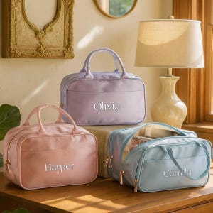 May include: Three rectangular travel bags with top handles and gold-tone zippers. The bags are pastel pink, lavender, and light blue. Each bag features a personalized name embroidered in white script: Harper on the pink, Olivia on the lavender, and Camila on the light blue bag.