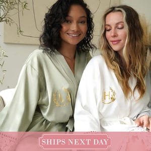 May include: A sage green robe and a white robe, both featuring long sleeves and a gold foil monogram inside a circular leaf wreath design on the chest. The robes have a smooth, satin-like finish and include matching waist ties for closure.