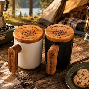 May include: Two ceramic mugs, one white and one black, each featuring a wooden handle engraved with the text 'Mrs. Garden' and a matching wooden lid. The lids are laser-engraved with a mountain landscape, a tent, a campfire, and the text 'The Adventure Begins Mr & Mrs. Garden'.