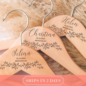May include: Three light-coloured wooden clothes hangers with silver metal hooks. Each hanger is personalised with a name, the date 05.24.2023, the word BRIDESMAID, and a black ink illustration of a delicate leafy branch across the bottom.
