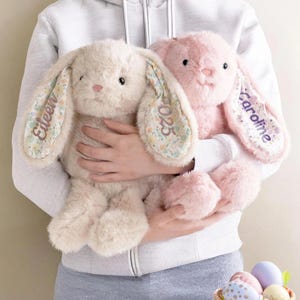 May include: Two plush stuffed bunnies, one cream and one pink, featuring long floppy ears with floral fabric linings. The cream bunny has the name Eileen and the year 2025 embroidered on its ears, while the pink bunny has the name Caroline embroidered on its ear in purple thread.