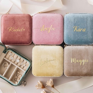 May include: Six square velvet jewellery cases in shades of pink, blue, and yellow, each with a gold name. One case is open, displaying rings and earrings. The cases are arranged on a light background.
