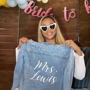 May include: A light blue denim jacket featuring white pearl embellishments along the upper back yoke and the embroidered white text "Mrs. Lewis" in a cursive script font centered on the back panel of the garment.