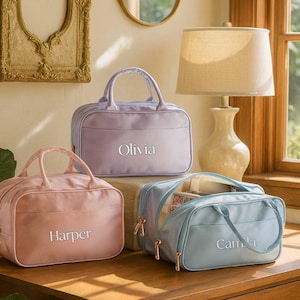 May include: Three pastel-coloured travel bags, pink, lavender, and blue, each with a top handle and zip closure. The bags are personalised with embroidered names. They are displayed on a wooden surface.