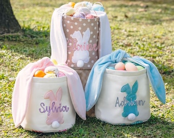 May include: Three fabric Easter baskets with long bunny ears. The back basket is tan with white polka dots and the name Eloise. The front left is white with a pink bunny and the name Sylvia. The front right is white with a blue bunny and the name Adrian.