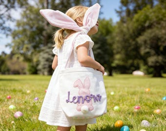 Può includere: Una borsa tote in tela bianca con una sagoma di coniglio in feltro rosa, una coda a pompon bianco e il nome Lenora ricamato in corsivo viola. La borsa ha manici lunghi ed è abbinata a un paio di lunghe e morbide orecchie da coniglio rosa.