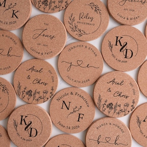 May include: A collection of round cork coasters with personalized text and designs. Each coaster features names, initials, and dates, with floral and leaf accents. Ideal for weddings or special events.