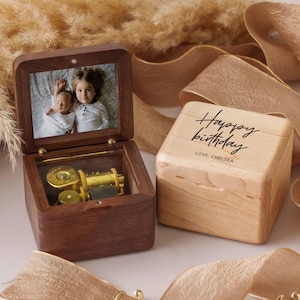 Peut inclure: Une boîte à musique en bois avec une photo à l'intérieur, à côté d'une boîte en bois clair avec "Happy birthday" et "Love, Chelsea" écrit dessus. La boîte à musique est ouverte, révélant un mécanisme musical doré.