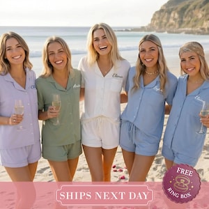 May include: Five matching pajama sets consisting of button-down shirts with collars and shorts in lavender, sage green, white, light blue, and dusty blue. Each shirt features a pocket with a name embroidered in cursive script on the chest.