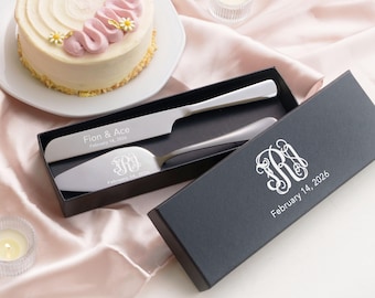 May include: A silver cake knife and server set in a black box. The knife is engraved with Fion & Ace and February 14, 2026. The server features an RA monogram and February 14, 2026. The box lid displays the same RA monogram and date in white text.
