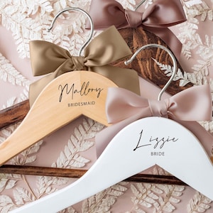 May include: Wooden and white hangers with satin bows. One hanger is engraved with "Mallory Bridesmaid", another with "Lizzie Bride". The bows are in shades of brown and pink. The hangers are on a pink and white floral background.