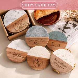 May include: A collection of round coasters featuring a split design of natural wood and stone. Each coaster is engraved with custom names, dates, and floral or monogram motifs. Some coasters are stored in a cardboard box or a gold metal holder.