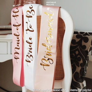 May include: Satin sashes in blush, white, and antique mauve, draped over a chair. The sashes are printed with gold lettering that reads "Maid of Honor", "Bride To Be", and "Bridesmaid". A decorative item for a bridal party.