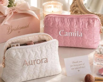May include: Two quilted cosmetic pouches, one white with the name Aurora embroidered in gold and one pink with the name Camila embroidered in white. Both feature a gold zipper pull and a soft, textured surface with a repeating heart pattern.