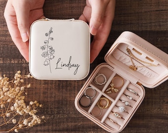May include: A square, cream-coloured jewellery travel case with a zip closure. The lid features a black line-art illustration of a lily of the valley flower and the name Lindsay. The interior is lined with light pink fabric and contains rings.