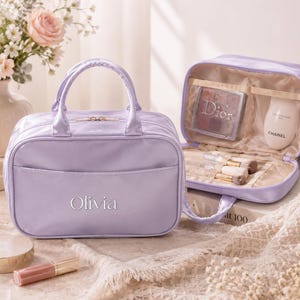 May include: A lavender cosmetic bag with two top handles and a zipper closure, featuring the name Olivia embroidered in white on the front. The open bag reveals internal pockets containing a Dior palette, a Chanel bottle, and makeup brushes.