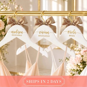 May include: Three white wooden hangers with gold metal hooks, each featuring a satin champagne-colored bow. The hangers are personalized with gold script text reading madison BRIDESMAID, vivian BRIDESMAID, and viola BRIDE, hanging on a gold rod.