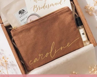 May include: A brown canvas zippered pouch with the name caroline in gold script, placed in a wooden gift box containing an Origins sheet mask, a small round tin, a Jo Malone glass spray bottle, and a card with the text Will you be my Bridesmaid?