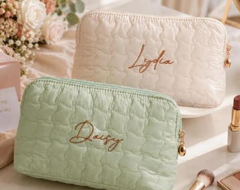 May include: Two rectangular quilted pouches with gold-tone zippers. The front pouch is light green with the name Daisy embroidered in brown script. The back pouch is cream with the name Lydia embroidered in brown script. Both have a soft texture.