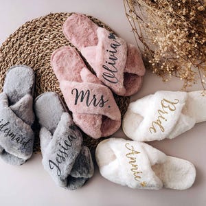 A collection of plush slippers with personalized names on them, arranged on a woven mat and accompanied by dried flowers.