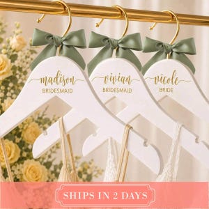 May include: Three white wooden clothes hangers with gold metal hooks, each adorned with a sage green satin bow. The hangers feature gold script text reading Madison Bridesmaid, Vivian Bridesmaid, and Nicole Bride, hanging on a gold rod.