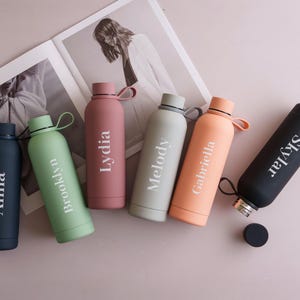 May include: Six matte finish water bottles in various colors including navy, sage green, dusty rose, light gray, peach, and black. Each bottle features a white printed name: Anna, Brooklyn, Lydia, Melody, Gabriella, and Skylar. Each has a matching cap.