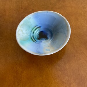 May include: A small, white ceramic bowl with a blue, green, and black swirl design. The bowl is glazed and has a glossy finish.