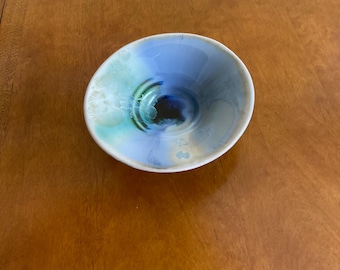May include: A small, white ceramic bowl with a blue, green, and black swirl design. The bowl is glazed and has a glossy finish.