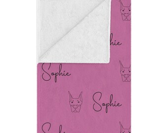 Puede incluir: Una manta rosa con un reverso blanco y esponjoso. La parte superior presenta un patrón repetido de la cara de un perro y el nombre "Sophie" en escritura negra. La manta está doblada, revelando el reverso blanco.