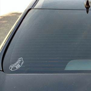 May include: A white vinyl sticker of a hot dog with the word "BOOF" underneath. The sticker is on the back window of a black car.