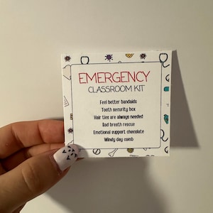 Pode incluir: Um cartão branco com o texto "EMERGENCY CLASSROOM KIT" em vermelho e preto. O cartão lista itens como "Feel better bandaids", "Tooth security box", "Hair ties are always needed", "Bad breath rescue", "Emotional support chocolate", e "Windy day comb".