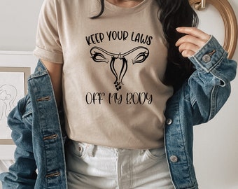 Peut inclure: Un t-shirt beige avec le texte "Keep your laws off my body" et une illustration noire d'un utérus.
