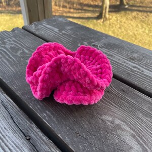 May include: A bright pink fuzzy hair scrunchie made of yarn. The scrunchie is on a wooden surface.