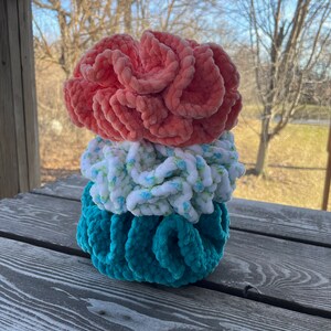 May include: Three crocheted hair scrunchies stacked on top of each other. The top scrunchie is coral pink, the middle scrunchie is white with a blue floral pattern, and the bottom scrunchie is teal.