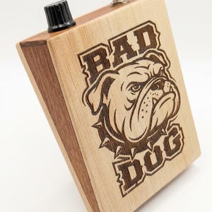 May include: A wooden box with a black knob and a silver button. The box features a laser-etched design of a bulldog wearing a spiked collar, with the words "BAD DOG" above and below the dog.