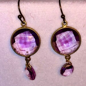 May include: A pair of dangle earrings with a round, purple, faceted gemstone set in a gold-toned metal frame. Each earring has a smaller, teardrop-shaped purple gemstone dangling from the bottom.