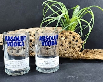 Könnte beinhalten: Zwei klare Glasbecher mit der Aufschrift "ABSOLUT VODKA" in Blau auf der Vorderseite. Die Becher stehen auf einem Stück Treibholz mit einer grünen Pflanze in einem braunen Topf.