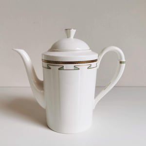 Könnte beinhalten: Weiße Kaffeekanne aus Keramik mit Deckel und Griff. Die Kanne hat ein gold-grünes geometrisches Design am oberen Rand. Der Ausguss ist lang und gebogen. Der Griff hat einen grünen Akzent. Auf dem Deckel befindet sich ein kleiner, dekorativer Knauf.
