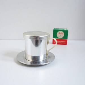 May include: A silver-colored Vietnamese coffee filter, with a matching saucer and lid. The filter has a clear plastic handle. A small green and red box is in the background.