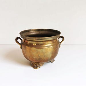 Puede incluir: Una jardinera de latón vintage con cuerpo redondeado, dos asas pequeñas y pies ornamentados. La jardinera tiene un aspecto deslustrado y envejecido, lo que sugiere que es antigua. El borde está ligeramente ensanchado y el interior es oscuro.