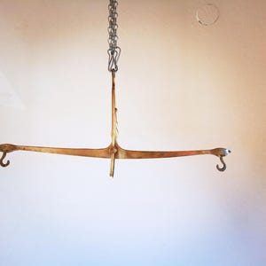 Op de afbeelding: Antieke messing balansweegschaal, opgehangen aan een ketting. De weegschaal heeft een horizontale balk met gebogen uiteinden en haken. De centrale steun is verticaal en verbonden met de ketting.