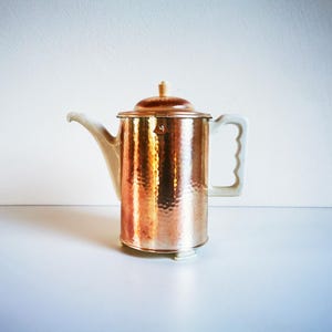 Op de afbeelding: Een koperkleurige koffiepot met een gehamerde textuur. Het heeft een witte handgreep en tuit, en een bijpassende deksel met een houten knop. De pot staat op een wit oppervlak, ideaal voor het serveren van koffie of thee. Hoogte: ongeveer 20 cm.