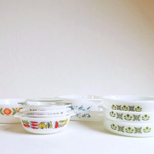 Op de afbeelding: Een verzameling vintage Pyrex-kommen in verschillende maten en patronen. De kommen hebben kleurrijke bloemen- en groentemotieven, met witte en transparante deksels. Sommige kommen zijn op elkaar gestapeld, waarbij de handvatten en decoratieve elementen zichtbaar zijn.
