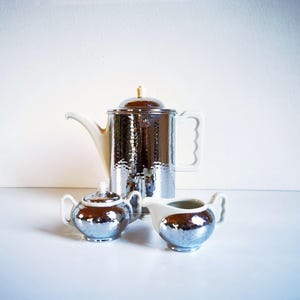 Könnte beinhalten: Ein silbernes Teeservice mit gehämmertem Muster, bestehend aus einer Teekanne, einer Zuckerdose und einem Sahnekännchen. Die Teekanne hat einen weißen Griff und Ausguss, und die Zuckerdose und das Sahnekännchen haben weiße Akzente. Das Set wird auf einer weißen Oberfläche präsentiert.