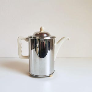 Op de afbeelding: Een vintage zilverkleurige koffiepot met een deksel, handvat en tuit. Het handvat en de tuit zijn gebroken wit. De pot heeft een cilindrisch lichaam en staat op kleine, gebroken witte voetjes.
