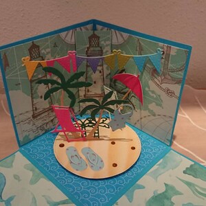 Könnte beinhalten: Eine 3D-Pop-up-Karte mit einer Strandszene. Die Karte zeigt Palmen, einen Strandstuhl, Flip-Flops, einen Sonnenschirm und eine Girlande mit bunten Flaggen. Die Karte ist aus Papier und hat ein blaues und grünes Farbschema.