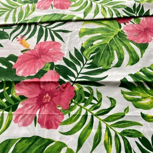 May include: White fabric printed with a tropical floral design. It showcases large pink hibiscus flowers, green monstera leaves, and palm fronds. This fabric is suitable for creating a summery, botanical-themed decor.