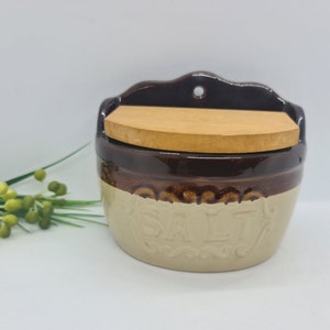 May include: A wall-mounted ceramic salt crock featuring a two-tone design with a dark brown glazed upper section and a light beige lower section. The word SALT is embossed on the front. It includes a flat, light-coloured wooden lid.